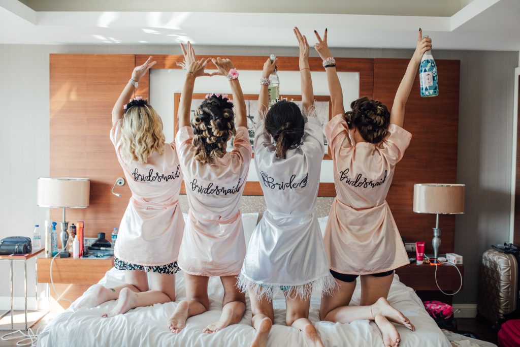 Four bridesmaids in robes celebrate in a hotel room, hands up for a fun pre-wedding party.